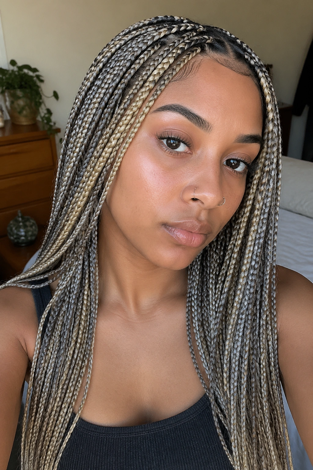 Close-up of a real woman with small knotless box braids in mixed blonde and grey, head-and-shoulders in a softly lit bedroom.